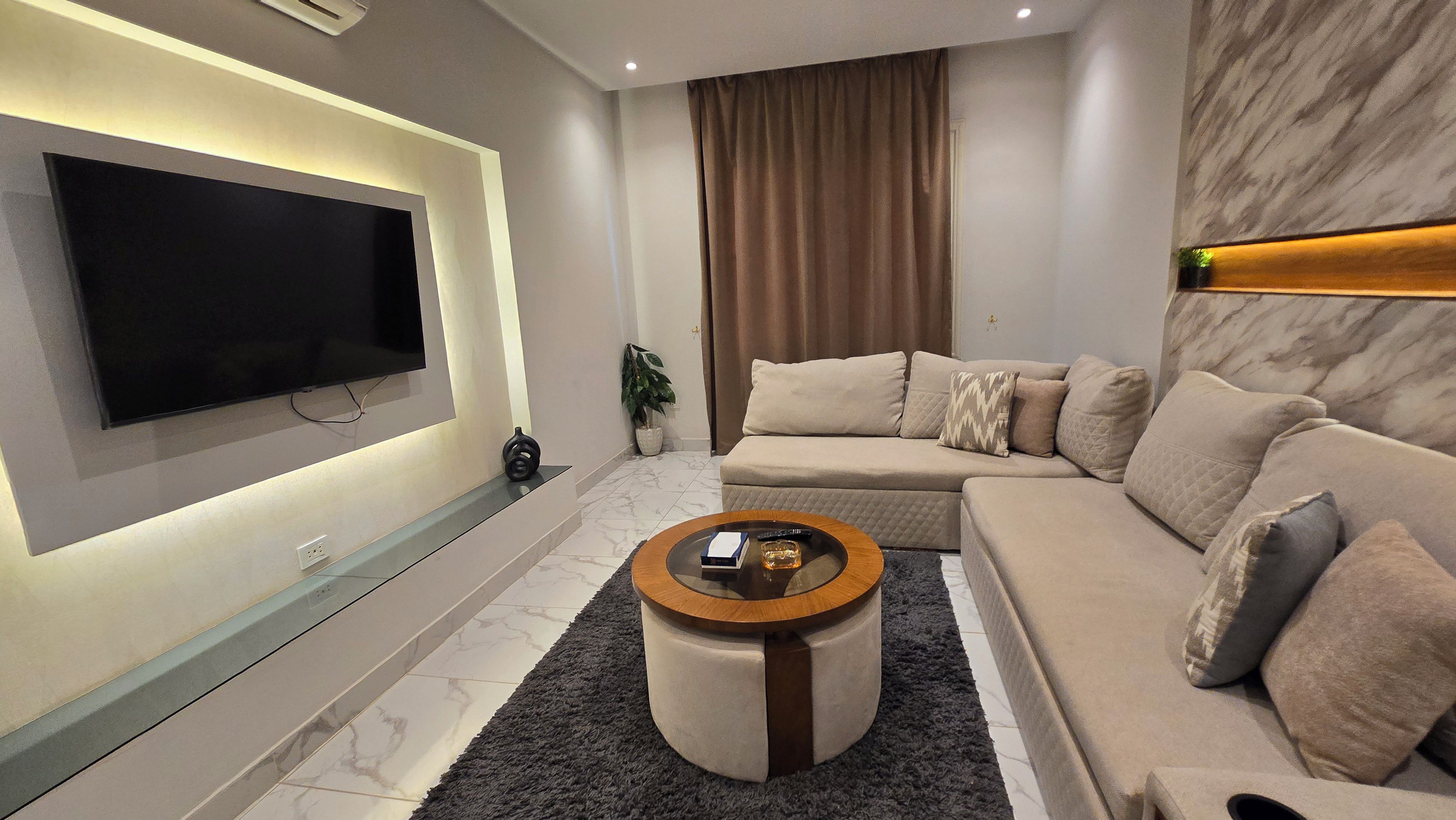 Un salon moderne avec un téléviseur à écran plat fixé au mur, un canapé beige, une table basse ronde et une plante en pot.