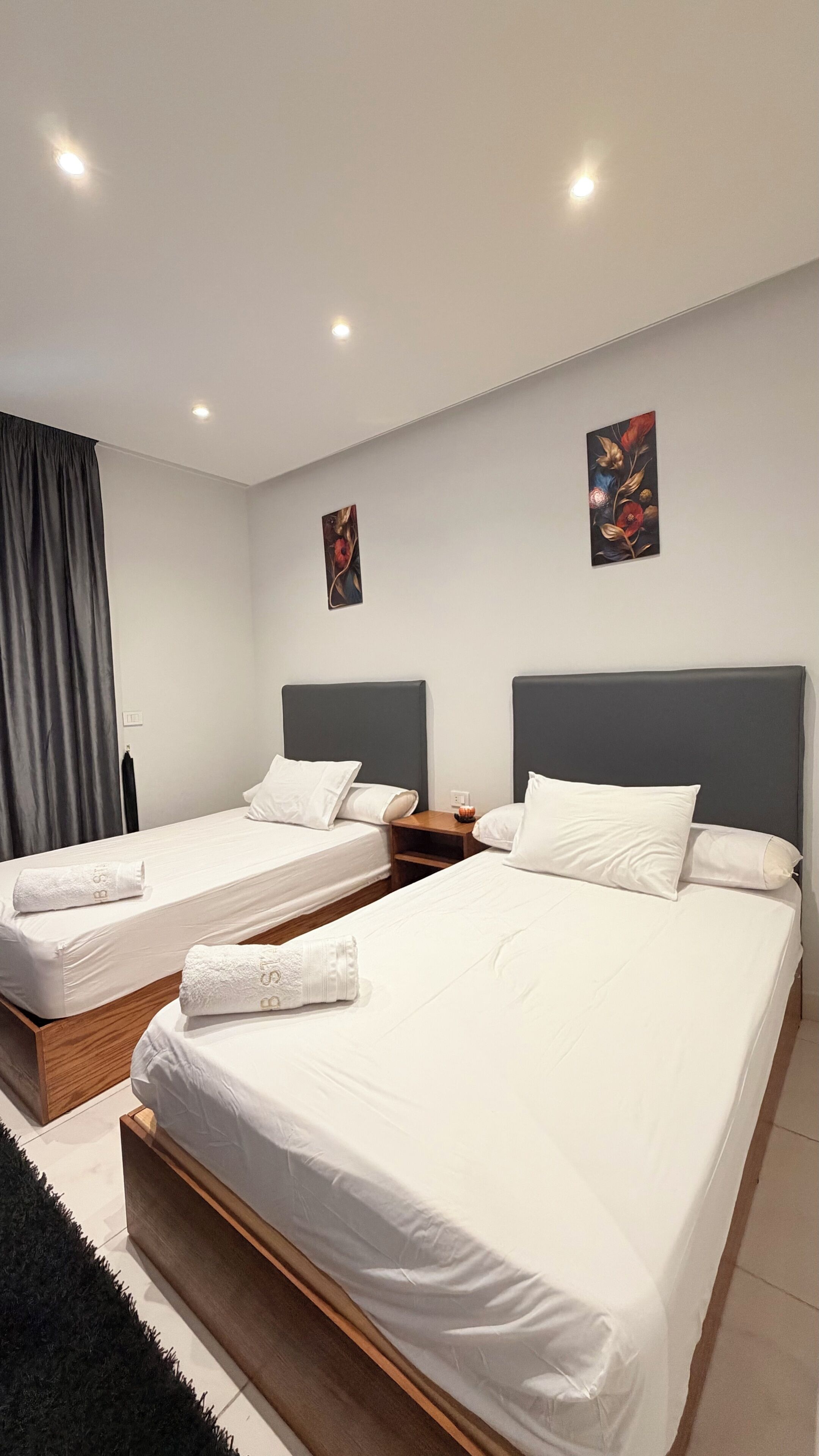 Hotellihuone, jossa on kaksi sänkyä, tumma verho, taulu seinällä ja pieni pöytä, jossa on lamppu.