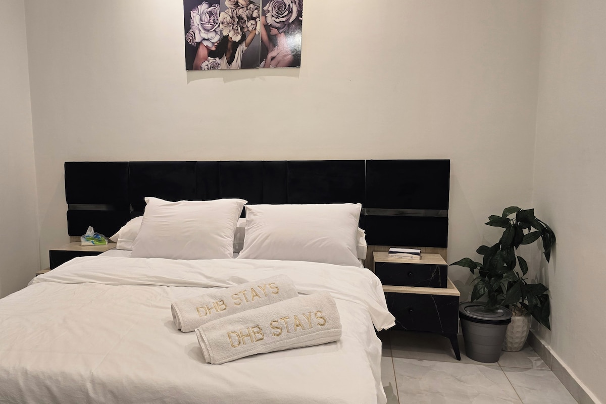 Een net opgemaakt bed met witte lakens en twee handdoeken met geborduurde tekst, een zwart hoofdbord, een nachtkastje met een boek en een potplant.