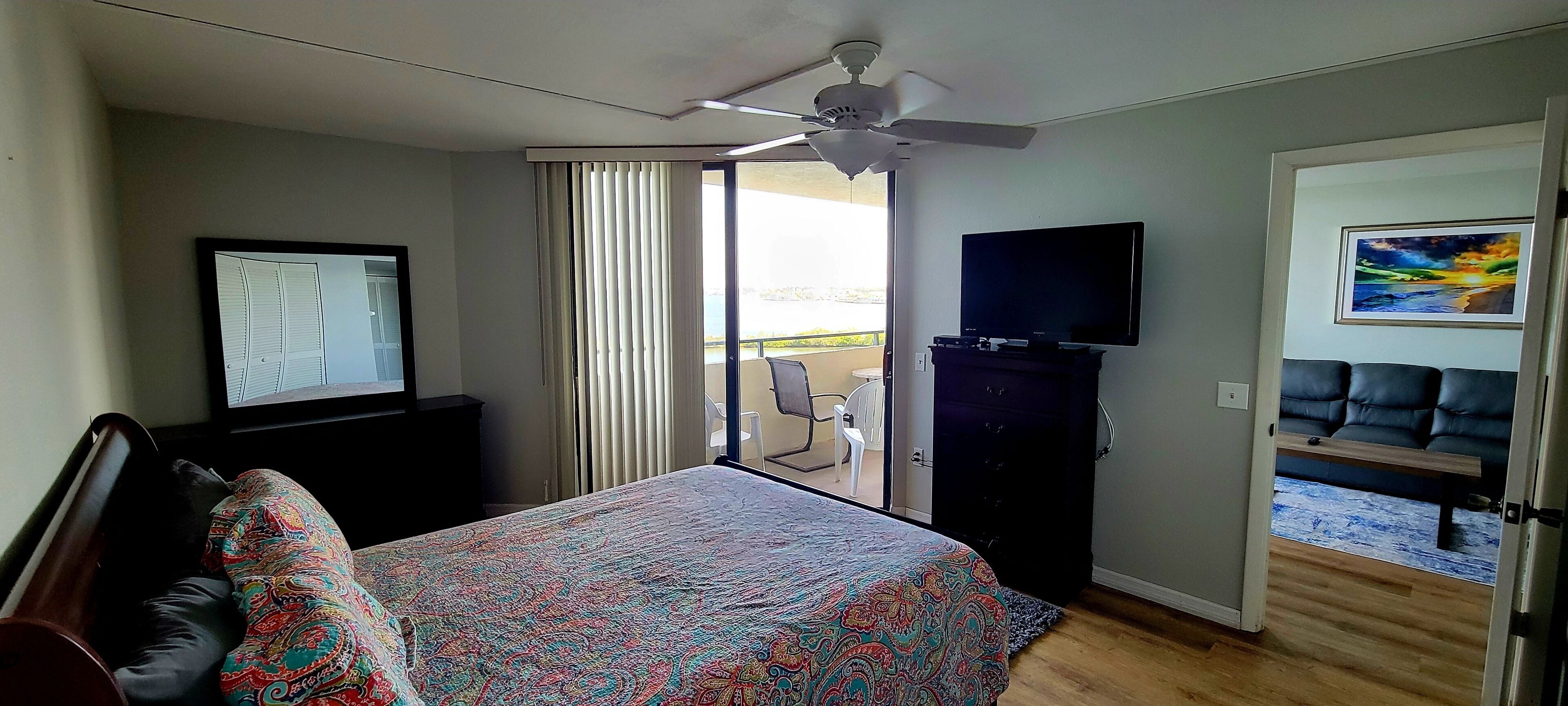 A hotel room with a bed, a TV, a ceiling fan, a balcony with a chair, and a painting on the wall.