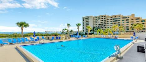 A swimming pool with blue chairs, palm trees, and a hotel building in the background.