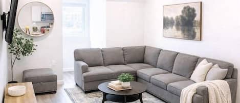 A modern living room with a grey sectional sofa, a round coffee table, and a flat-screen TV mounted on the wall.