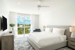 A modern hotel room with a large bed, a ceiling fan, a TV, a bedside table, and a view of the outdoors. - Ellipse Resort, Grace Bay /Jr Suite One Bedroom with Grace Bay Street View (Grace Bay)