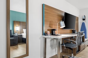 Moderni hotellihuone, jossa on suuri peili, työpöytä lampun kanssa, tuoli, sänky ja kahvinkeitin.