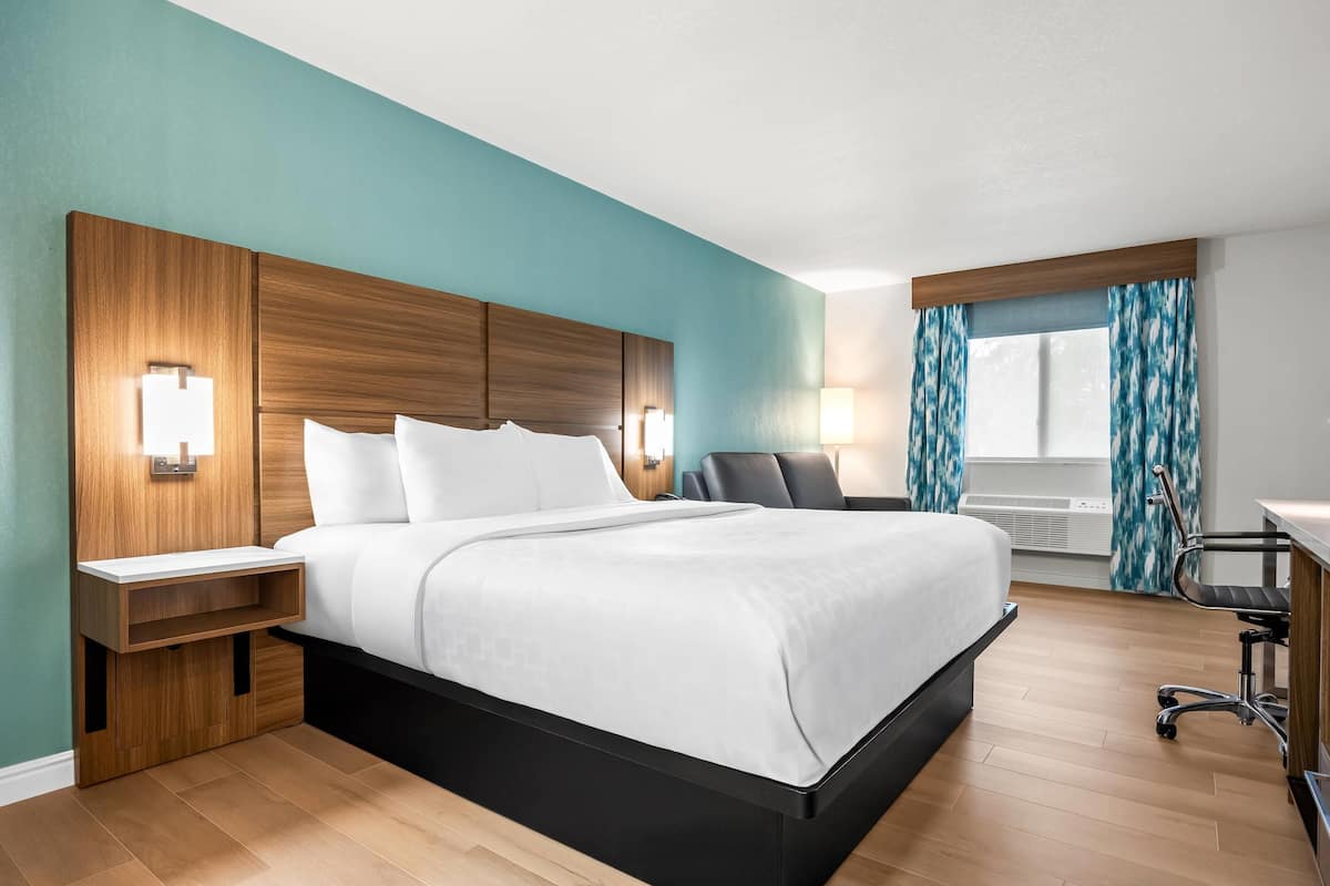 A modern hotel room with a wooden headboard, a bed with white linens, a desk with a chair, and a window with curtains.