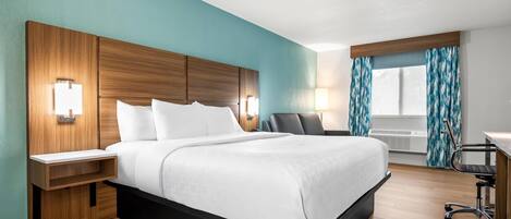 A modern hotel room with a wooden headboard, a bed with white linens, a desk with a chair, and a window with curtains.