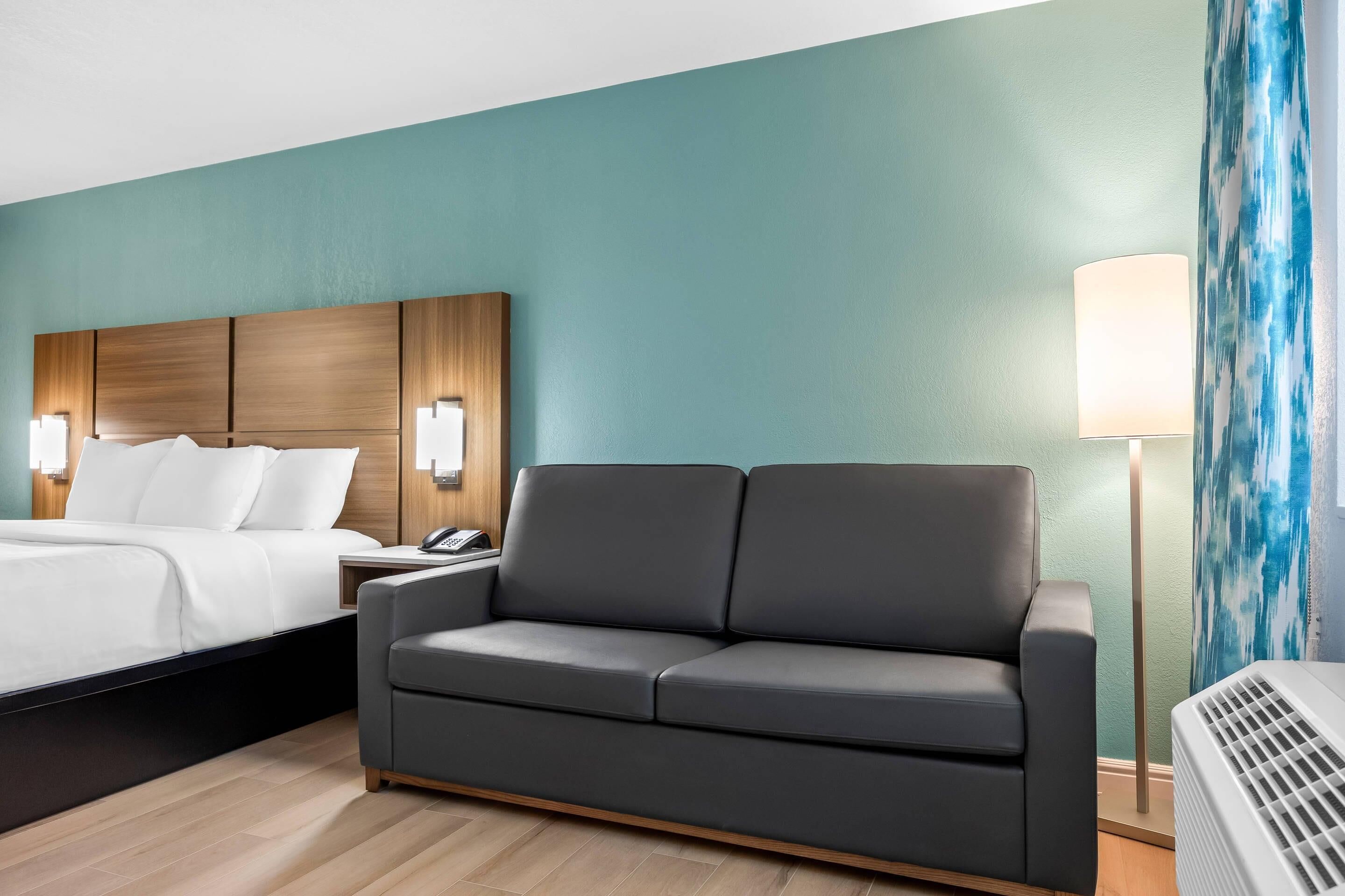 A hotel room with a bed, a gray sofa, a nightstand with a phone, a lamp, and a wall with a blue abstract painting.