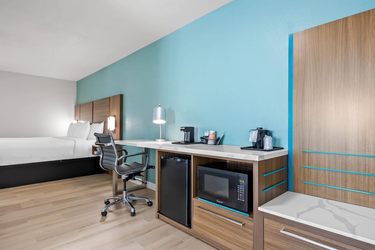 A hotel room with a bed, desk, refrigerator, microwave, and a chair.