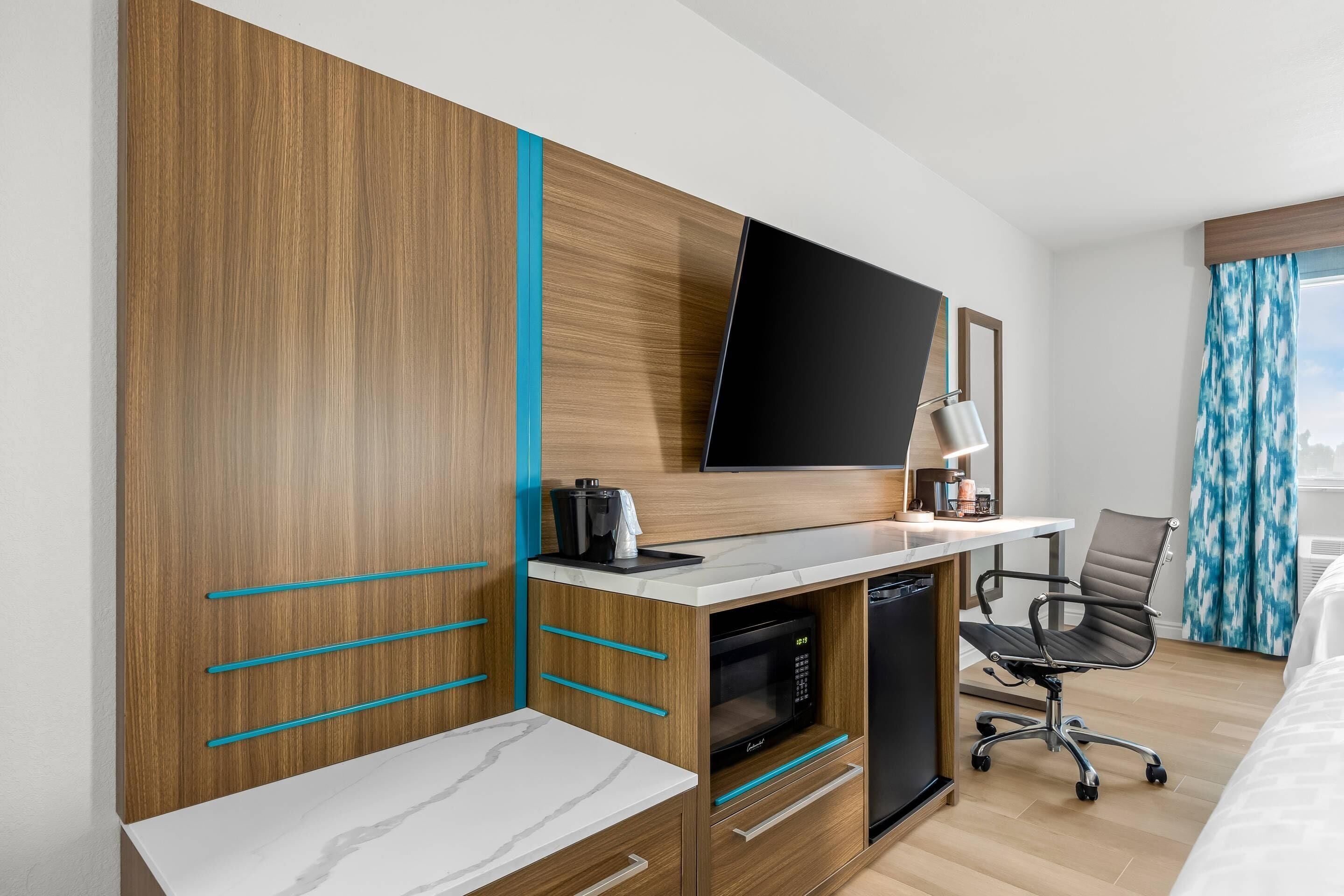 A hotel room with a wooden wardrobe, a flat-screen TV, a desk with a microwave, a chair, and a bed.