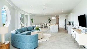 Ein modernes Wohnzimmer mit einem blauen, gebogenen Sofa, einem Essbereich, einer Küche und einem großen Fernseher rechts.