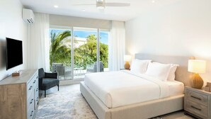 Ein modernes Hotelzimmer mit einem großen Bett, einem Flachbildfernseher, einem Schreibtisch mit Stuhl und einem Balkon mit Blick auf Palmen und den Himmel.