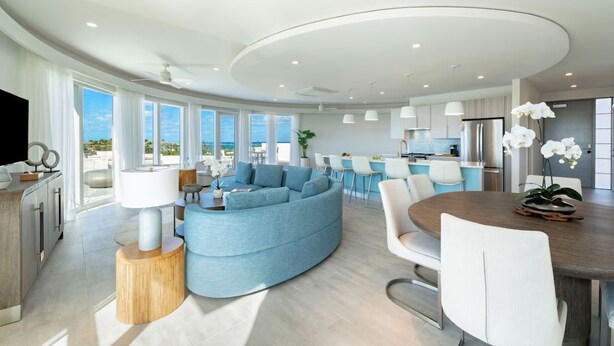 A modern living room with a curved sofa, a dining table, and a kitchen area with a bar. - Ellipse Resort, Grace Bay/Family Suite Two Bedroom with Private Terrace (Grace Bay)