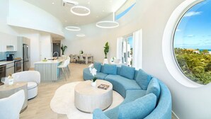 A modern interior with a curved sofa, a round coffee table, and a dining area with a kitchen in the background. - Ellipse Resort, Grace Bay/Family Suite Two Bedroom with Private Terrace (Grace Bay)