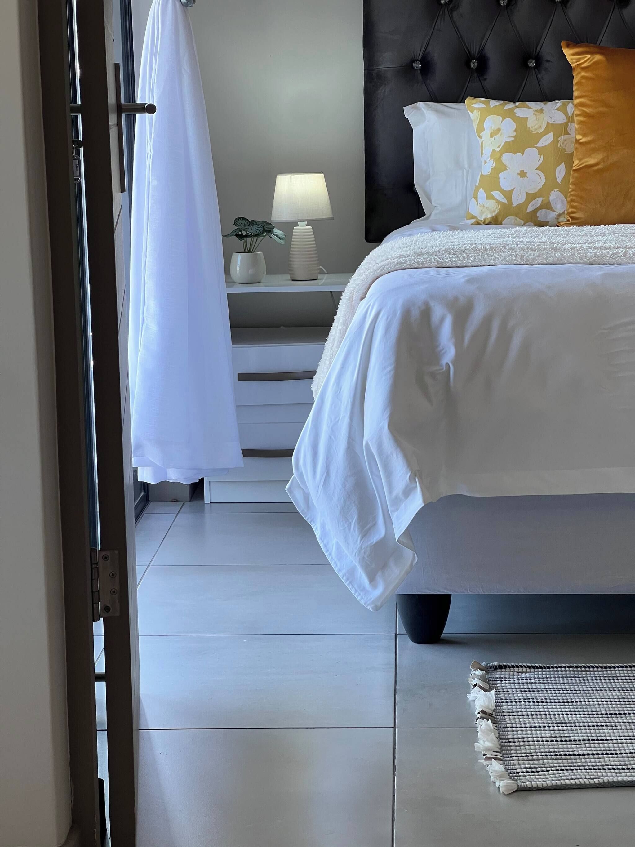 A bedroom with a bed, bedside table, lamp, and a view of the outside.