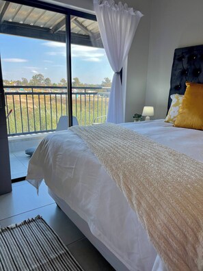 A bedroom with a large bed, a nightstand, a lamp, and a view of a balcony with a railing. - Amazing 1BR With River View in Waterfall-midrand (Johannesburg)