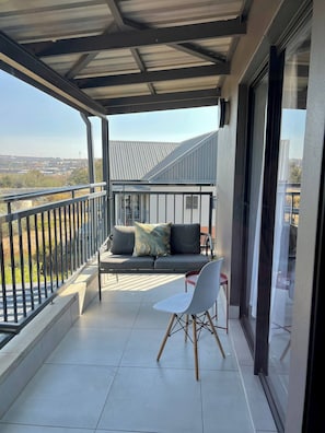 A balcony with a sofa and a chair, a metal railing, and a view of the cityscape. - Amazing 1BR With River View in Waterfall-midrand (Johannesburg)