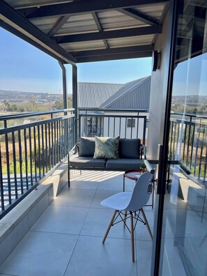 A balcony with a sofa, a chair, and a glass railing overlooking a landscape. - Amazing 1BR With River View in Waterfall-midrand (Johannesburg)