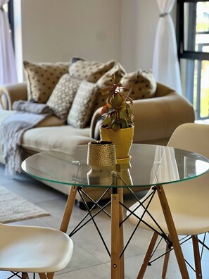 A living room with a glass table, two chairs, a sofa with patterned cushions, and a potted plant. - Amazing 1BR With River View in Waterfall-midrand (Johannesburg)
