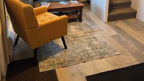 Ein Wohnzimmer mit einer Couch, einem gelben Sessel, einem Couchtisch, einem Fenster, einer Treppe und einem an der Wand hängenden Bild.