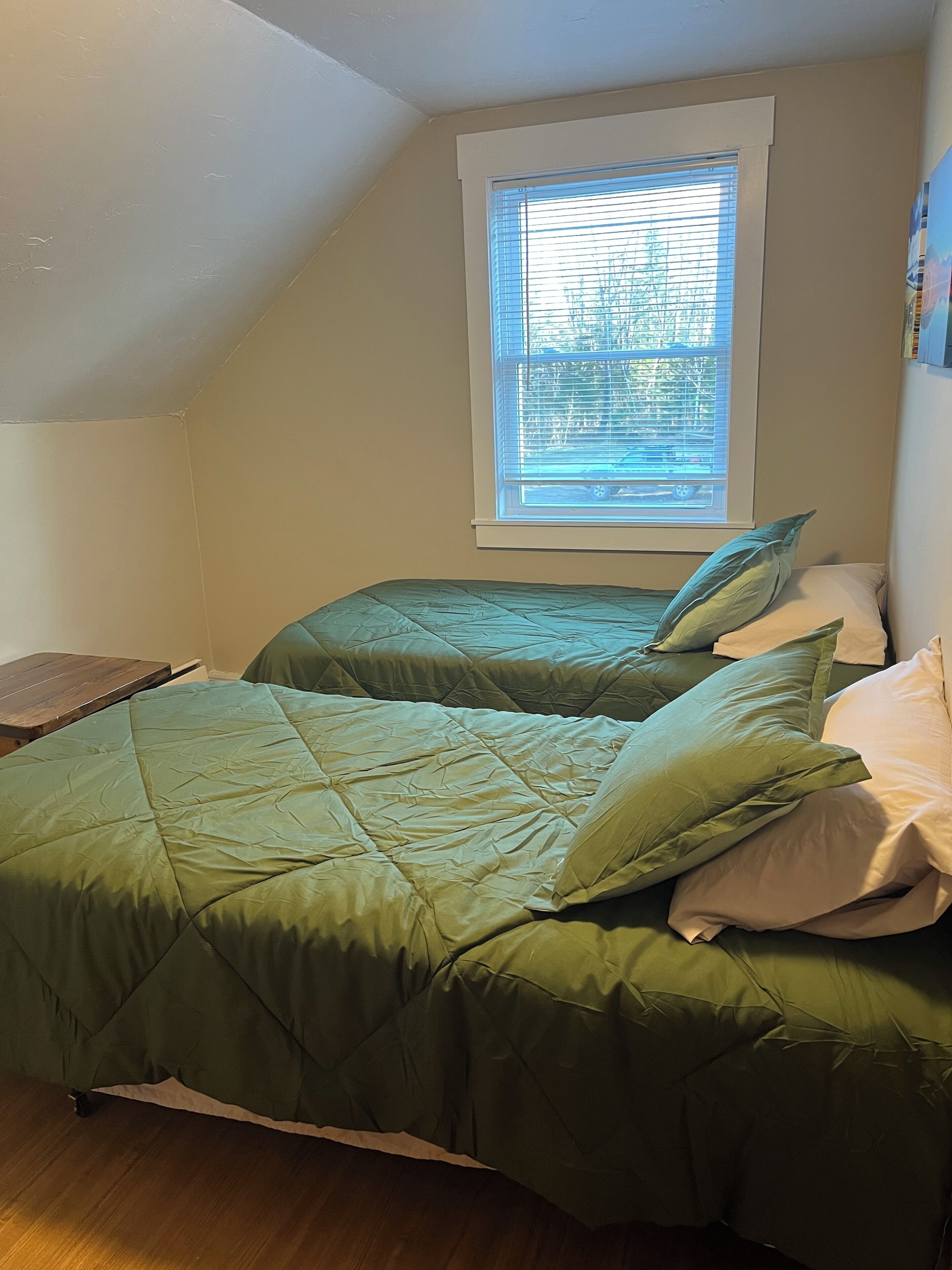 Ein kleines Schlafzimmer mit nur einem Raum, geneigter Decke, Fenster mit Jalousien, zwei Betten mit grüner Bettwäsche und einem Nachttisch aus Holz.