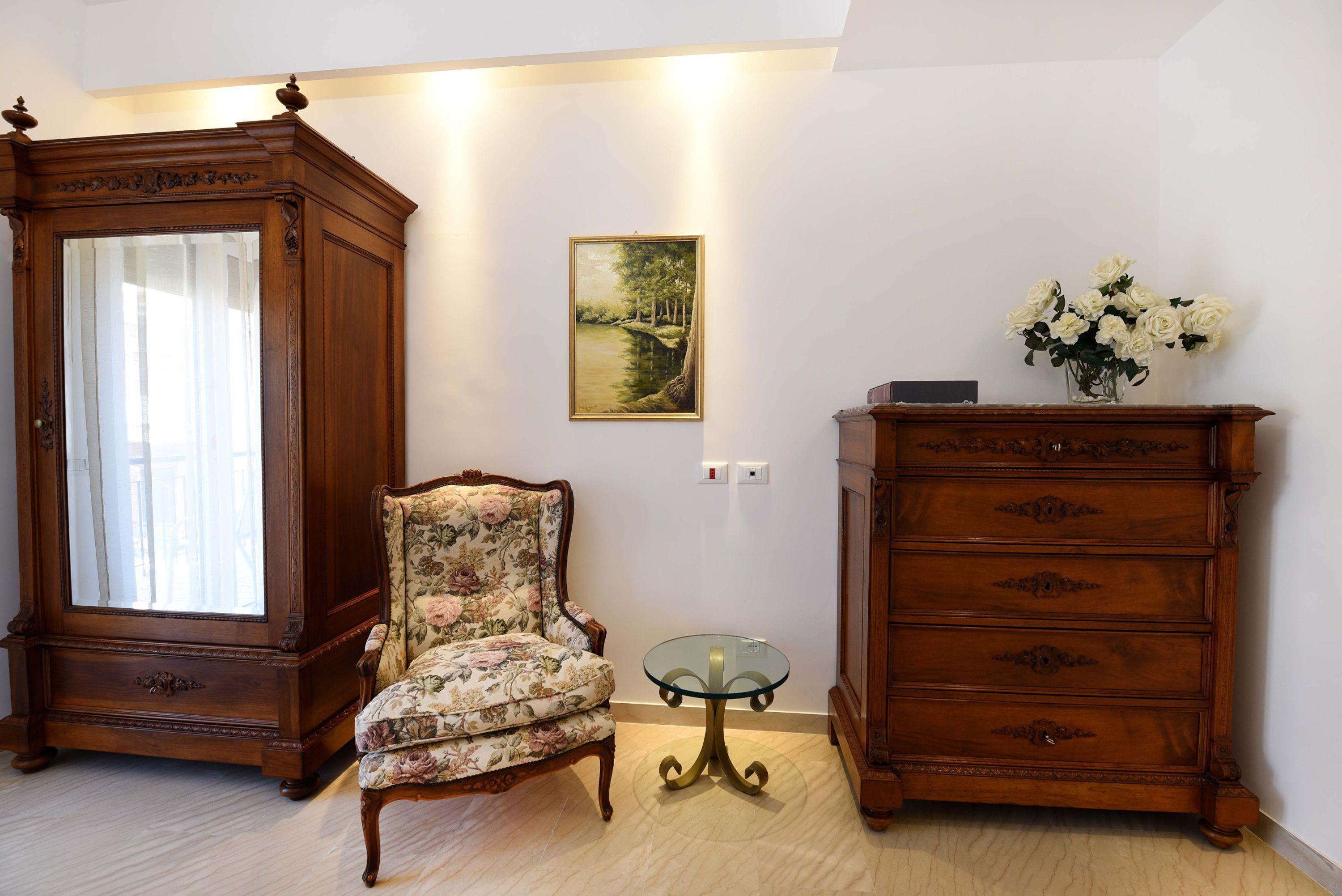 Quarto com um grande armário de madeira, uma poltrona floral, uma mesa de vidro e um quadro na parede.