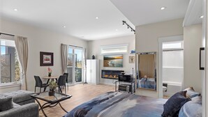 A modern hotel room with a large bed, a seating area with a glass table, a fireplace, and a balcony with a view.