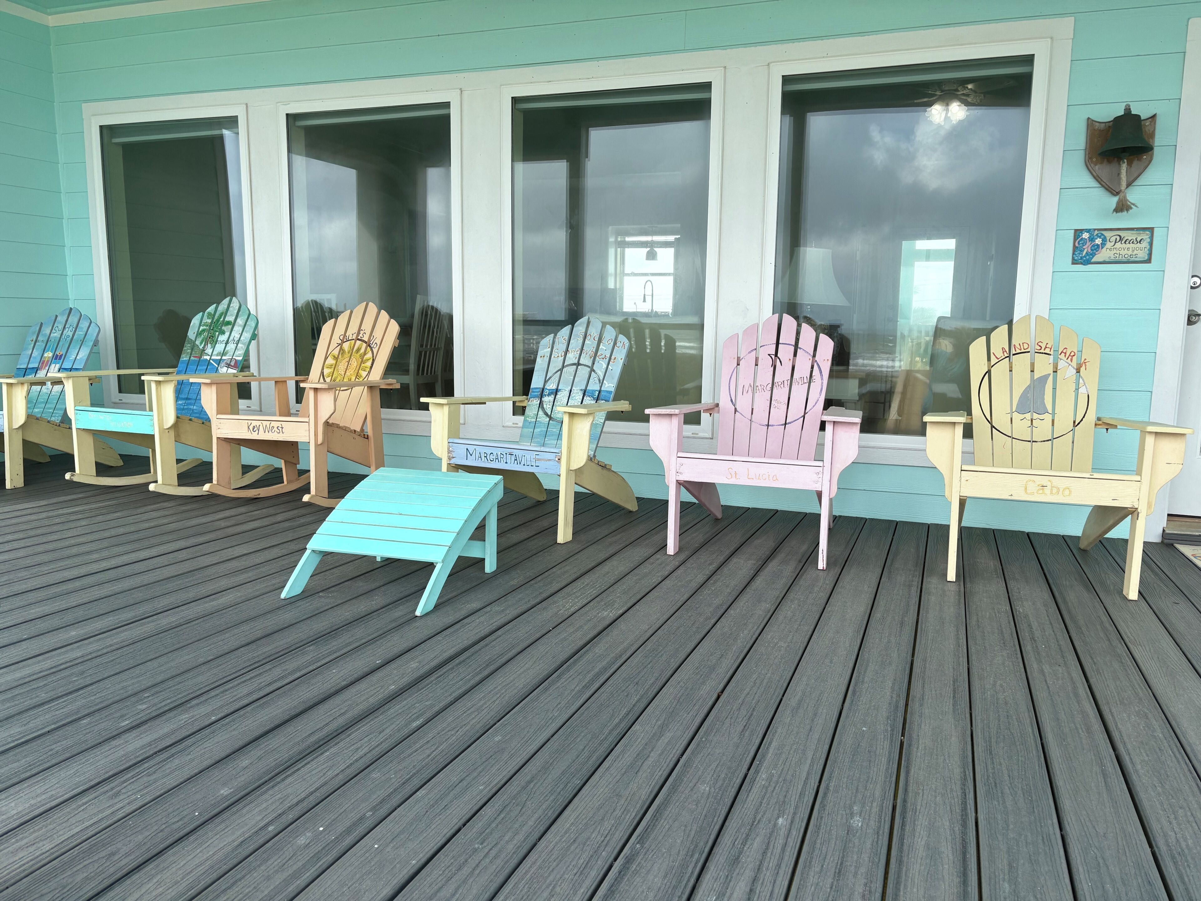 A row of Adirondack chairs on a wooden deck with a teal wall and windows in the background.
