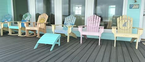 A row of Adirondack chairs on a wooden deck with a teal wall and windows in the background.