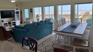 A spacious living room with a large blue sofa, a dining table with chairs, a TV, and a view of the beach through large windows. - Beachfront Escape – Beach Views from Wraparound Porch•Private Walkover W Seating (Freeport)