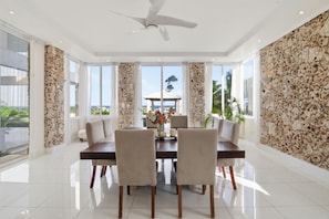 Un comedor con una mesa, sillas, un ventilador de techo y ventanas grandes con vistas al exterior.