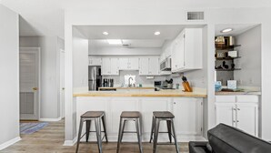 A modern kitchen with white cabinetry, a central island with stools, and a black leather couch in the adjacent living area. - Coastal Shores - Direct Oceanfront w/ Pool in North Myrtle Beach (North Myrtle Beach)