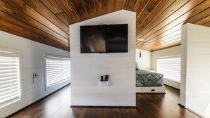 Une pièce avec un plafond en bois, un téléviseur fixé au mur, un lit recouvert d’une courtepointe à motifs verts et un meuble de rangement blanc.