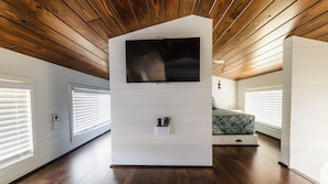 Une pièce avec un plafond en bois, un téléviseur fixé au mur, un lit recouvert d’une courtepointe à motifs verts et un meuble de rangement blanc.