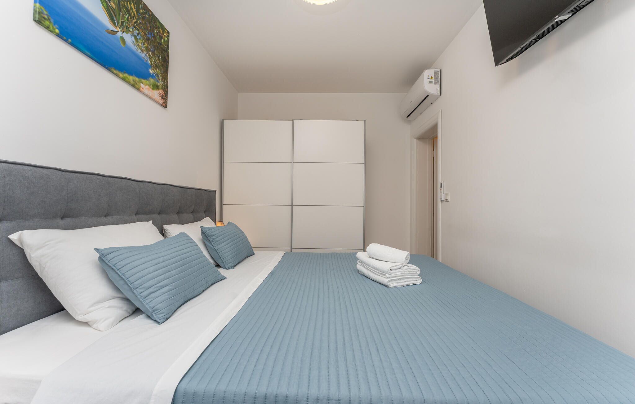 Quarto com cama, almofadas, colcha azul, televisão, aparelho de ar condicionado e guarda-roupa.