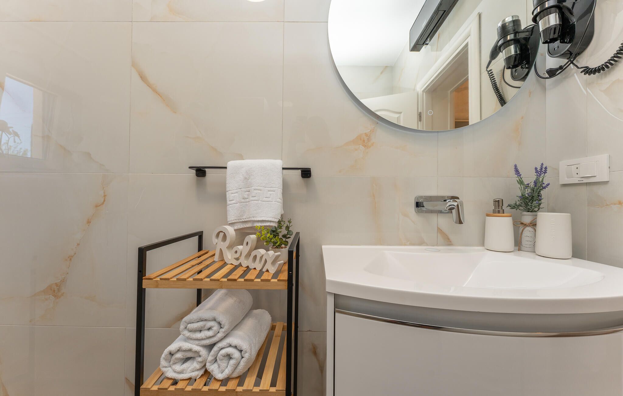 Ein Badezimmer mit marmorverkleideter Wand, einem runden Spiegel, einem Waschbecken und einem Handtuchhalter mit ordentlich aufgerollten Handtüchern.