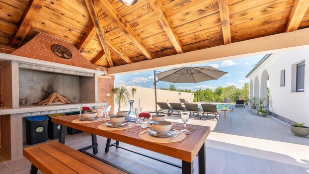 Patio esterno con tavolo in legno apparecchiato per mangiare, barbecue e area piscina sullo sfondo.