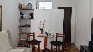 Ein kleiner Raum mit einer Couch, zwei Stühlen, einem Tisch mit Blumen und einem Bücherregal sowie einer Uhr.