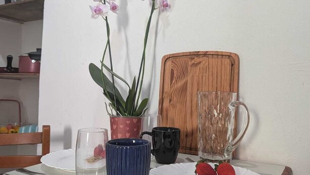 Ein Esstisch mit einer Schale Erdbeeren, einer blauen Vase mit Blumen, einem Glas und einem Holzschneidebrett.