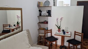 Ein kleiner, gut beleuchteter Raum mit einem weißen Sofa, einem Esstisch mit Stühlen, einem Spiegel, Regalen mit Büchern und Küchenutensilien sowie einer Zimmerpflanze.