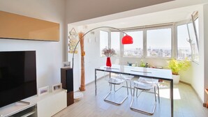 Ein modernes Wohnzimmer mit einem Flachbildfernseher, einem Essbereich mit einem Glastisch und Stühlen sowie einem großen Fenster mit Blick auf die Stadt.
