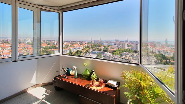 Ein Zimmer mit großem Fenster und Blick auf die Stadt, einem Holztisch mit verschiedenen Gegenständen und einer Topfpflanze.