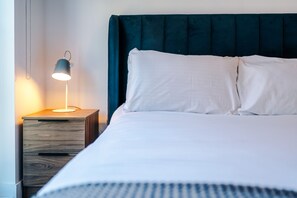 Ein ordentlich bezogenes Bett mit weißen Textilien, einem dunkelblauen Kopfteil und einem Nachttisch mit Lampe.