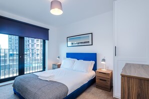 Ett modernt hotellrum med en stor säng, ett sängbord, en liten avlastningsyta, ett fönster med utsikt över byggnader och en inramad tavla på väggen.