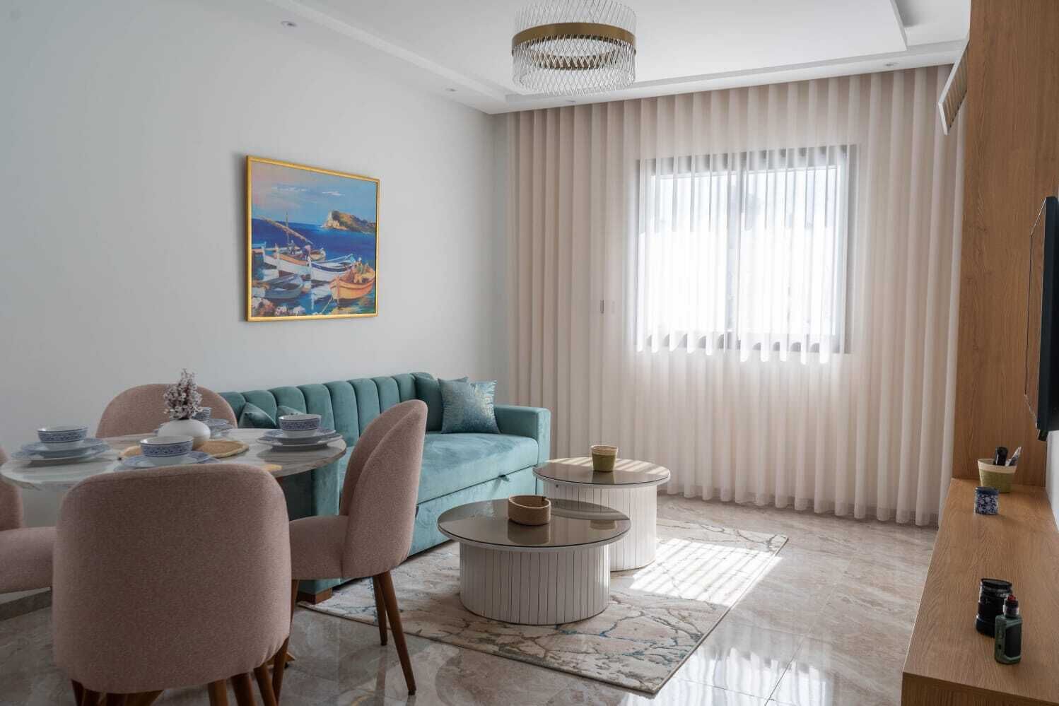 A modern living room with a teal sofa, a dining table set with blue and white plates, and a painting of boats on the wall.