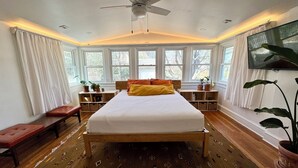 A bedroom with a bed, a ceiling fan, a TV, a window with curtains, a bedside table, and a plant. - Sunny, safe, walkable house with King bed and AC (Kansas City)