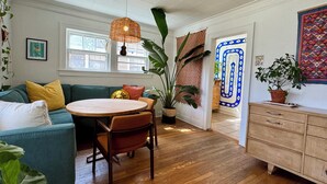 A cozy living room with a green sofa, wooden dining table, and chairs, a large plant, and a patterned door. - Sunny, safe, walkable house with King bed and AC (Kansas City)