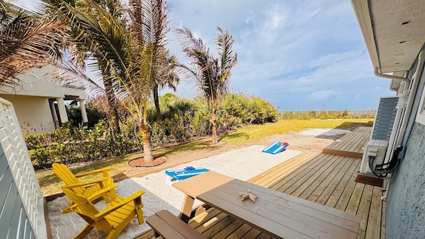 A wooden deck with a table and chairs, a covered area with a blue mat, and a view of the beach and palm trees. - BEACHHOUSE #1 Mermaids Paradise-full kitchen, private oceanfront patio 1st floor (Melbourne Beach)