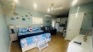 A bedroom with a bed, nightstands, a ceiling fan, a small white table, and a kitchen area with cabinets and appliances. - BEACHHOUSE #1 Mermaids Paradise-full kitchen, private oceanfront patio 1st floor (Melbourne Beach)