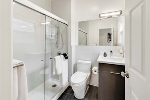 Un baño moderno con ducha acristalada, inodoro, lavabo con espejo y suelo de baldosas oscuras.