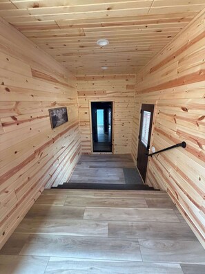 Interior de madera con una escalera central, una puerta a la derecha y un cuadro en la pared izquierda.